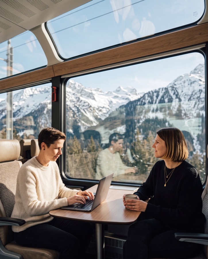 KI-Bild: Franka arbeitet am Laptop, Kathi trinkt Kaffee, im Panoramazug durch die verschneiten Alpen